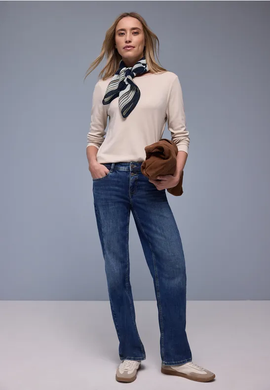 Femme en pull beige, jean bleu, foulard, sac à main, posée devant un fond neutre.