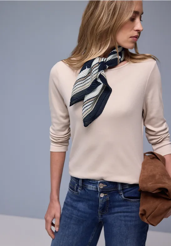 Femme portant un haut beige, un jean bleu, un foulard rayé et tenant une veste marron.