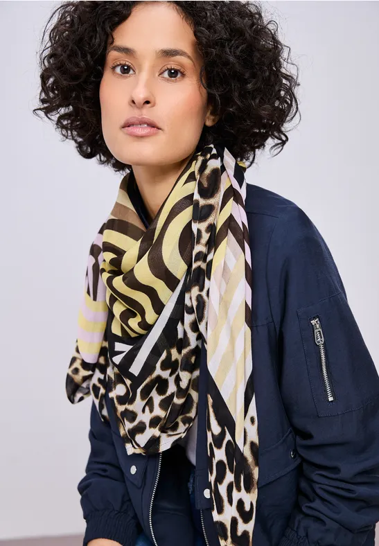 Een persoon met krullend haar draagt een marineblauw bomberjack en een kleurrijke sjaal met zebra- en luipaardprint.