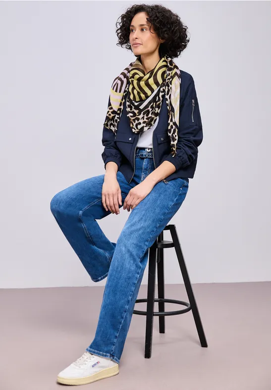 Persoon op een kruk draagt blauwe jeans, wit T-shirt, donkermarineblauwe jas, gemêleerde sjaal en witte sneakers.