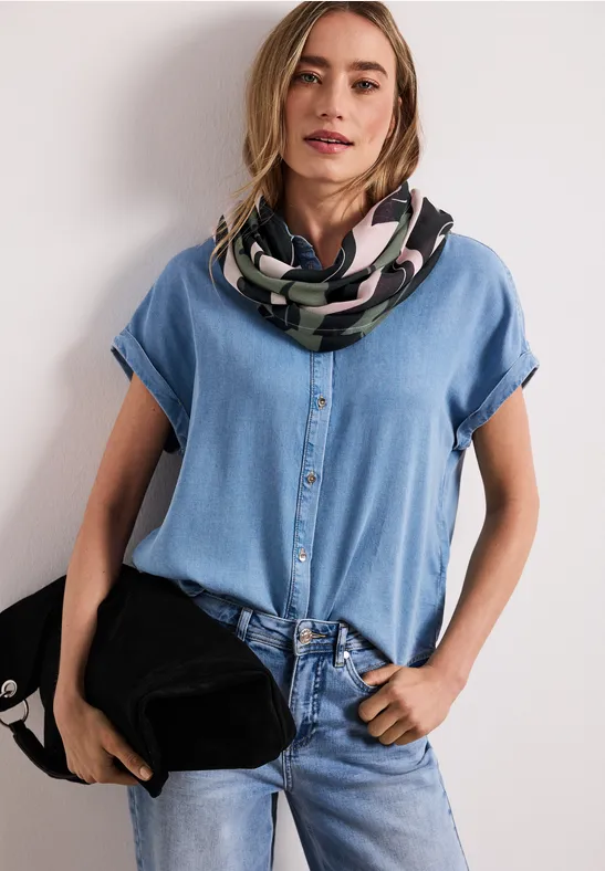 Vrouw in spijkerblouse en jeans met een patroon sjaal, houdt een zwarte tas vast.