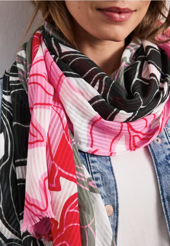 Vrouw met een roze-zwarte, patroonige sjaal en spijkerjasje voor een witte muur.
