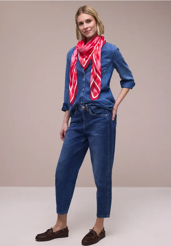 Frau trägt Jeanshemd, Jeanshose und rotes Halstuch, leger kombiniert mit braunen Slippern vor neutralem Hintergrund.