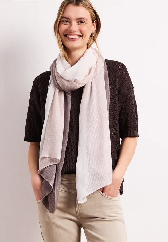 Frau mit hellem Schal, schwarzem Cardigan und beiger Hose, Hände in den Taschen.