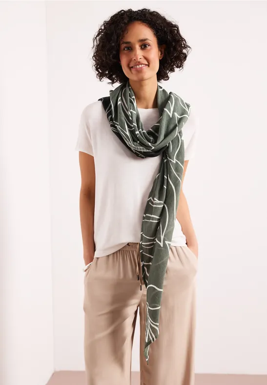 Une personne porte un t-shirt blanc, un pantalon beige et une écharpe verte autour du cou.