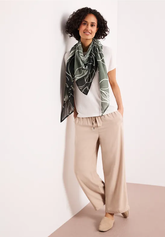 Personne avec foulard vert à motifs, t-shirt blanc et pantalon ample beige, appuyée contre un mur.