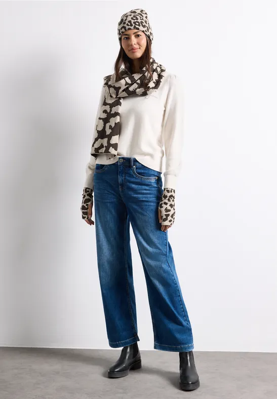 Frau mit weißem Pullover, blauen Jeans, schwarzen Stiefeletten und Leopardenmuster-Set aus Mütze, Schal und Handschuhen.