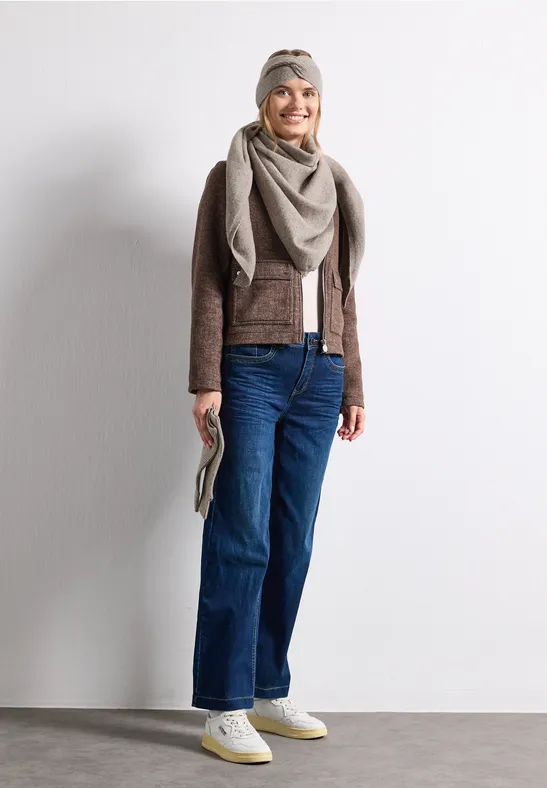 Vrouw voor lichte muur, lacht, draagt jeans, bruine jas, sjaal, hoofddoek, witte sneakers en clutch.