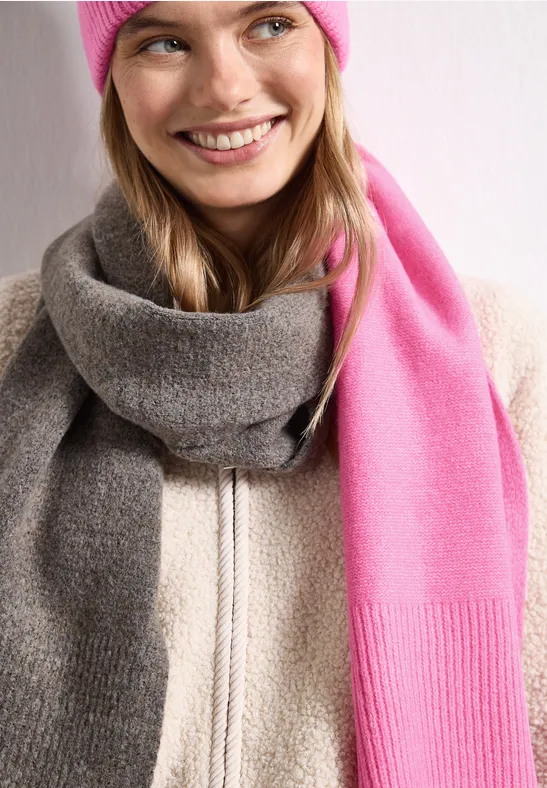 Jonge vrouw lacht, draagt een roze muts, een lange tweekleurige sjaal (roze en grijs) en een lichte jas.