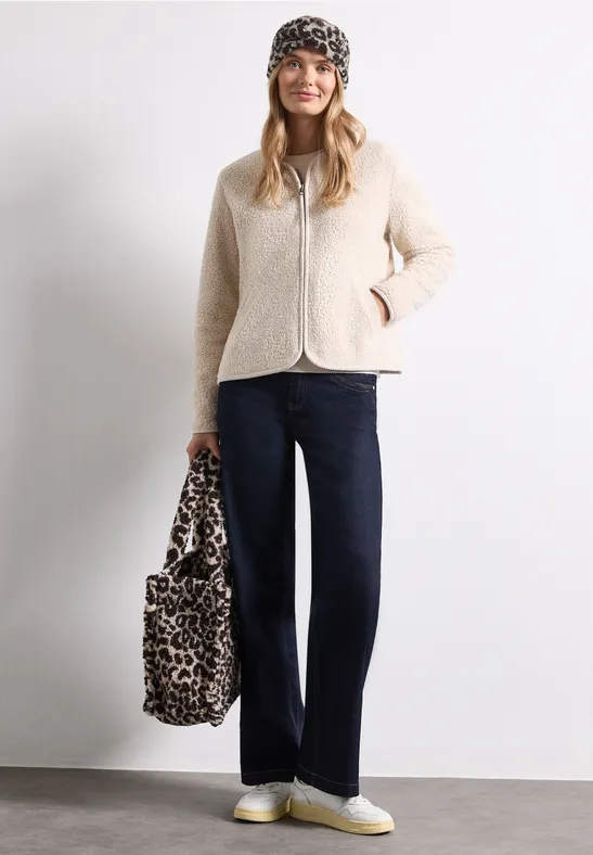 Vrouw met beige teddyjas, zwarte muts, donkere jeans, witte sneakers en luipaardprinttas voor een witte muur.