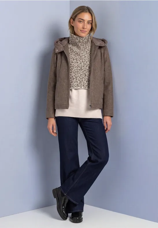 Eine Frau trägt eine braune Kapuzenjacke, cremefarbenen Pullover, weite Jeans und schwarze Schuhe vor hellem Hintergrund.