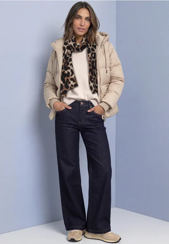 Frau in beiger Steppjacke, weißem Pullover, Leopardschal, weiten Jeans und Sneakers vor hellblauem Hintergrund.