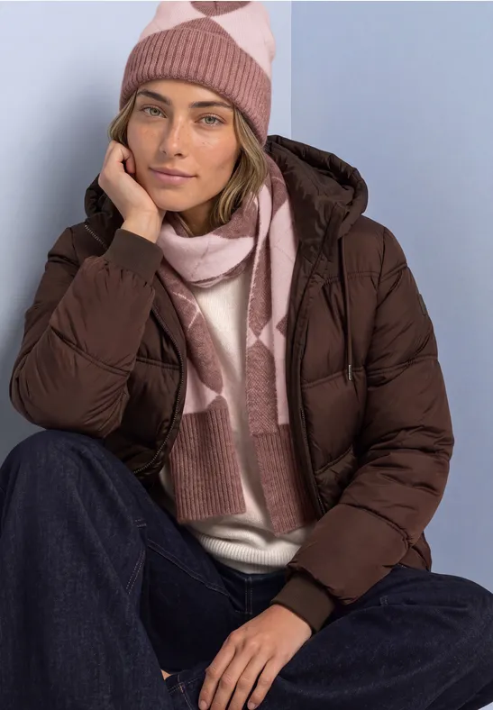 Eine Frau in brauner Steppjacke, Jeans, rosa-beigem Strickmütze und Schal sitzt und stützt den Kopf auf die Hand.