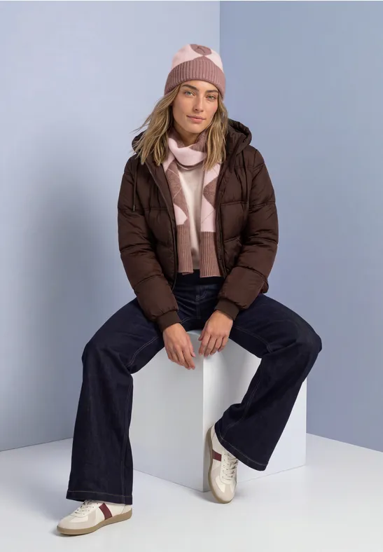 Frau sitzt auf Würfel, trägt braune Steppjacke, rosa-weiß gestreifte Mütze und Schal, dunkle Jeans, weiße Sneakers.