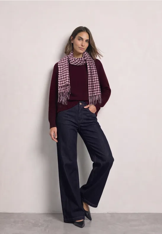 Frau in weinrotem Pullover, weitem Jeans, Hahnentritt-Schal und schwarzen Pumps vor hellem Hintergrund.
