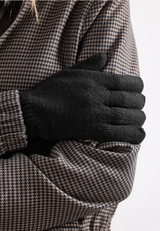 Nahaufnahme: Person in Hahnentrittmantel mit schwarzen Handschuhen, die Arme verschränkt vor der Brust.