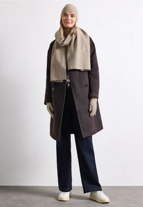 Een vrouw draagt een winterjas, sjaal, muts, handschoenen en broek in neutrale kleuren.