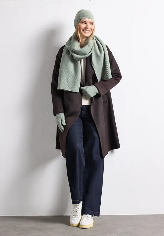 Vrouw lacht in grijze muts, sjaal en handschoenen, zwarte mantel, blauwe jeans en witte sneakers.