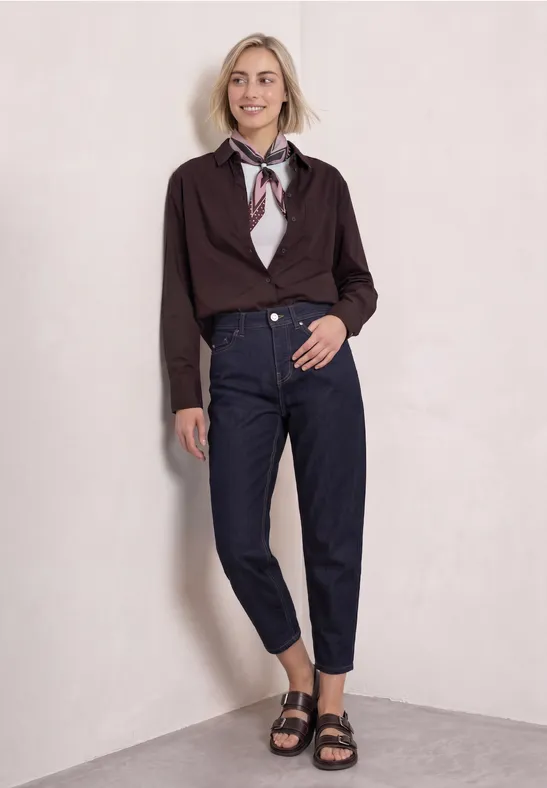 Junge Frau lehnt lächelnd an heller Wand, trägt dunkelblaue Jeans, Knotenbluse, Halstuch und schwarze Sandalen.