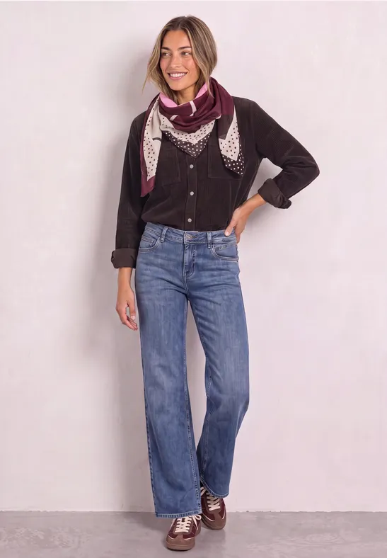 Frau mit Halstuch, schwarzem Pullover, blauen Jeans und Loafers lächelt vor hellem Hintergrund.