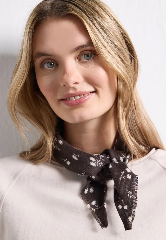 Jonge vrouw met blond haar draagt licht shirt en gebloemd sjaaltje om haar nek, ze glimlacht zacht.