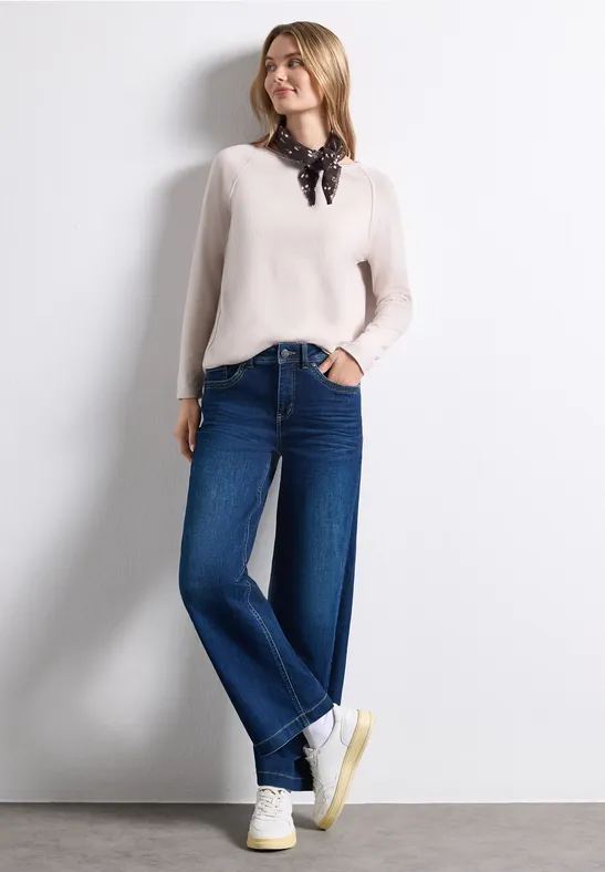 Een vrouw in een witte blouse, zwarte sjaal, wijde jeans en witte sneakers voor een lichte achtergrond.