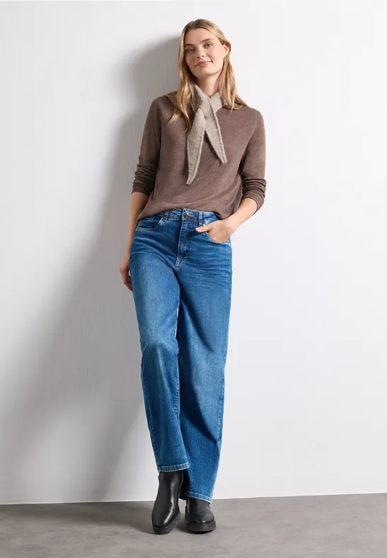 Frau steht vor weißer Wand, trägt braunen Pullover, Halstuch, weite blaue Jeans und schwarze Stiefeletten.