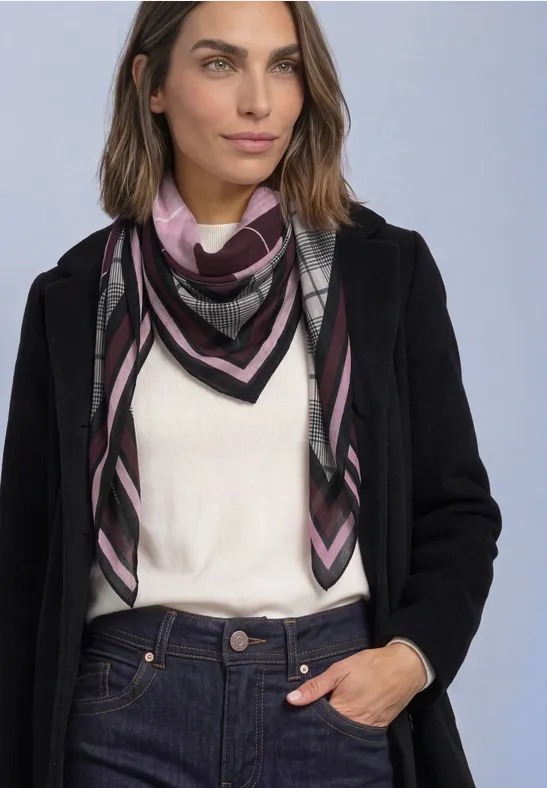 Femme en manteau noir, haut blanc, jean et foulard à motif rose et noir sur fond neutre.