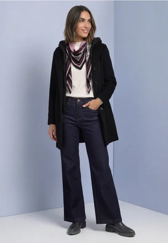Femme en manteau noir, chemisier clair, foulard et jean évasé, debout et souriante sur fond clair.