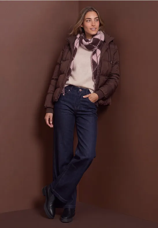 Femme en doudoune marron, pull blanc, jean et écharpe devant un fond brun.