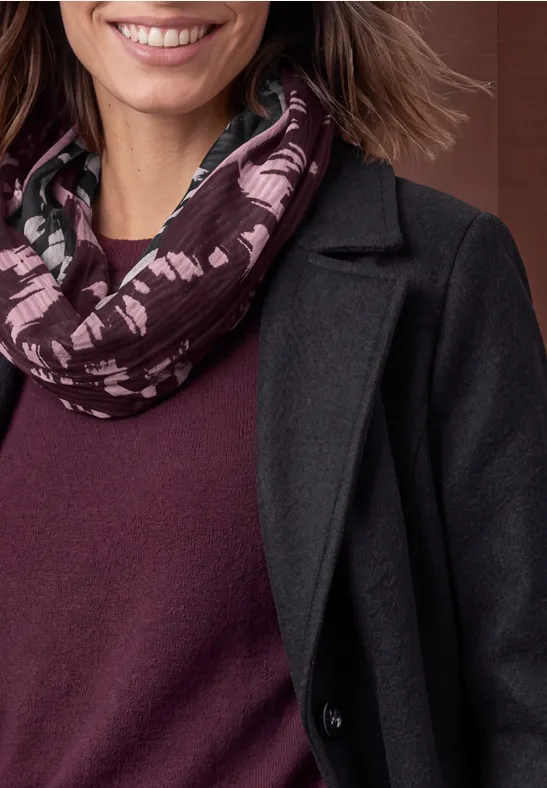 Gros plan sur une femme souriante en manteau gris foncé, pull bordeaux et écharpe à motifs.