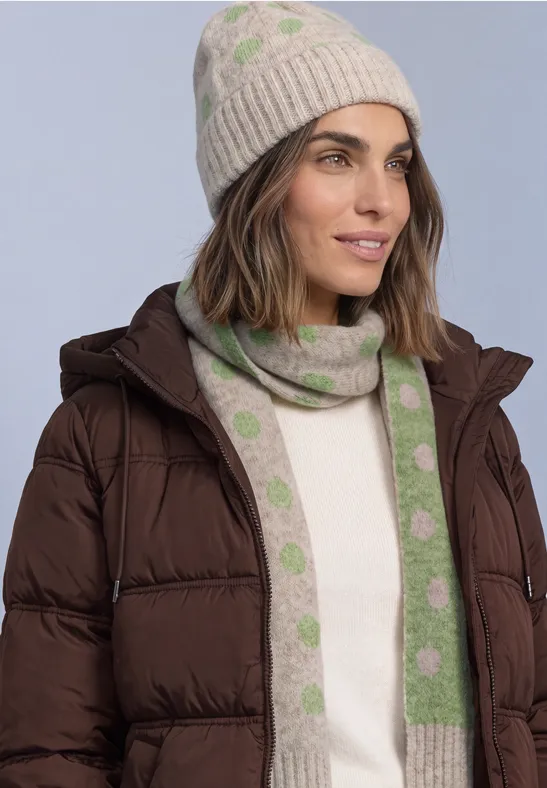 Frau in brauner Daunenjacke mit beigem Pullover, gestrickter Mütze und passendem Schal mit grünen Punkten.