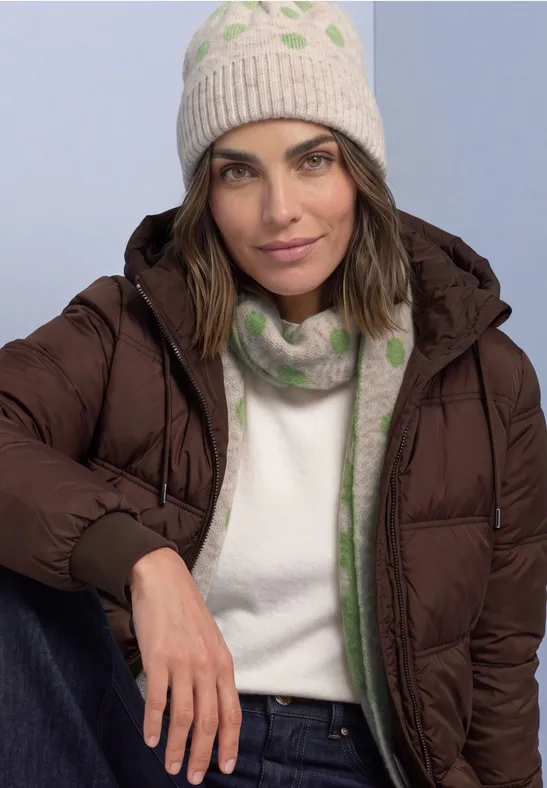 Frau in brauner Daunenjacke, cremefarbener Strickmütze und grün gemusterter Wollschal, lässig lehnend.