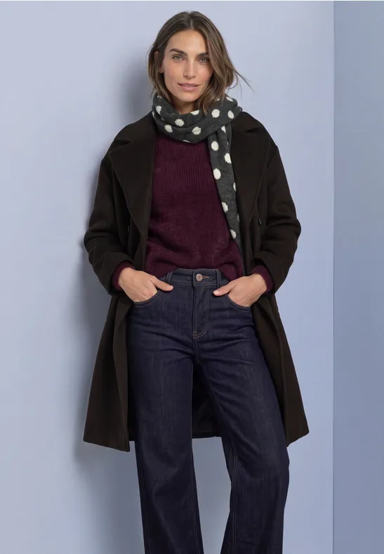 Frau in schwarzem Mantel, gepunktetem Schal, weinrotem Pullover und dunkler Jeans vor hellem Hintergrund.