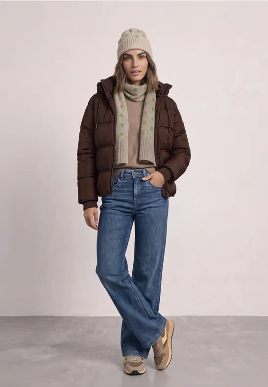 Femme portant une doudoune marron, jean, bonnet et écharpe beiges, posant devant un fond clair.