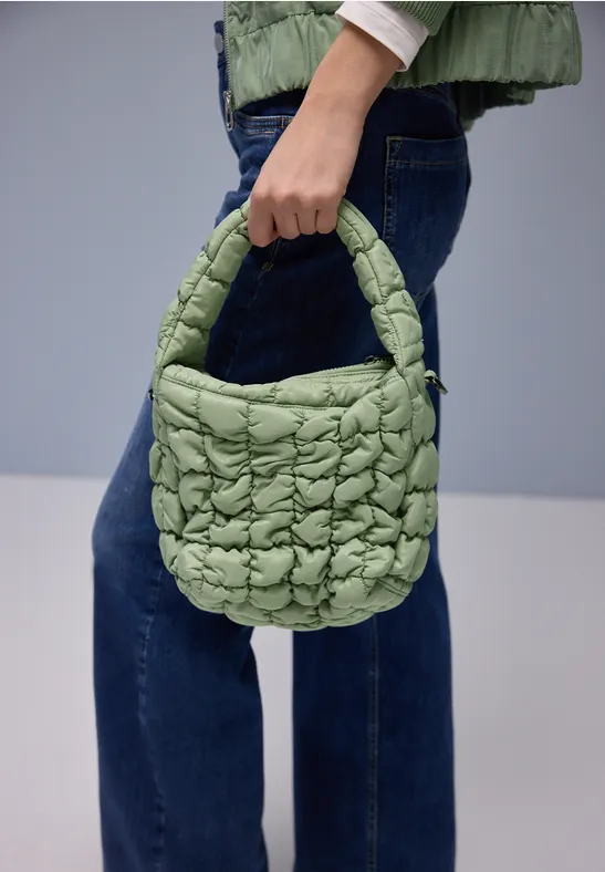 Eine Hand hält eine grüne, gesteppt gepolsterte Umhängetasche vor blauen Jeans.