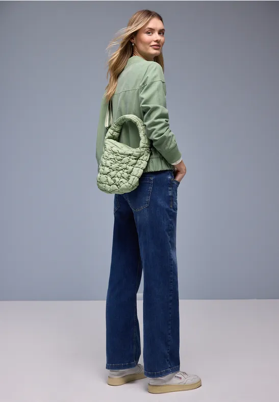 Eine Frau in grüner Steppjacke, weiten Jeans, weißen Sneakers und passender gesteppter Tasche vor grauem Hintergrund.