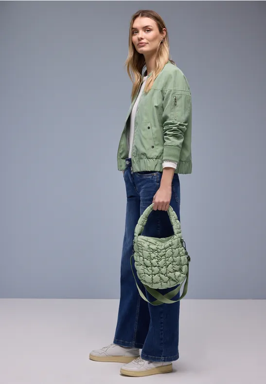 Person trägt grüne Jacke, weite Jeans und hält eine geflochtene grüne Tasche.