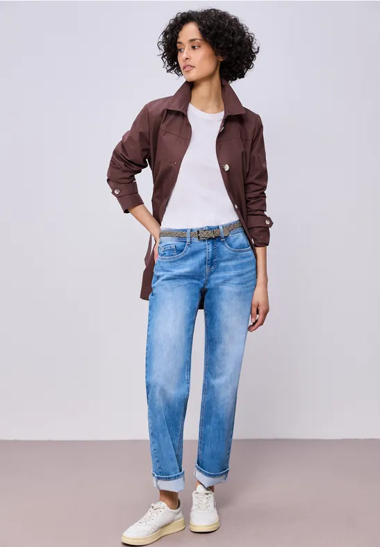 Een vrouw draagt een wit t-shirt, een bruine jas, blauwe jeans en witte sneakers voor een effen achtergrond.