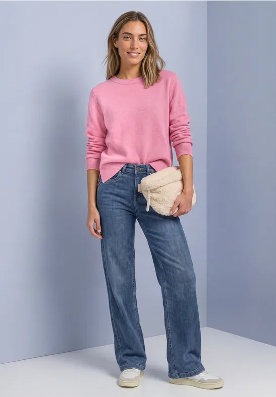 Eine Frau trägt rosa Pullover, weite Jeans, weiße Sneaker und hält eine beige Gürteltasche vor hellgrauer Wand.