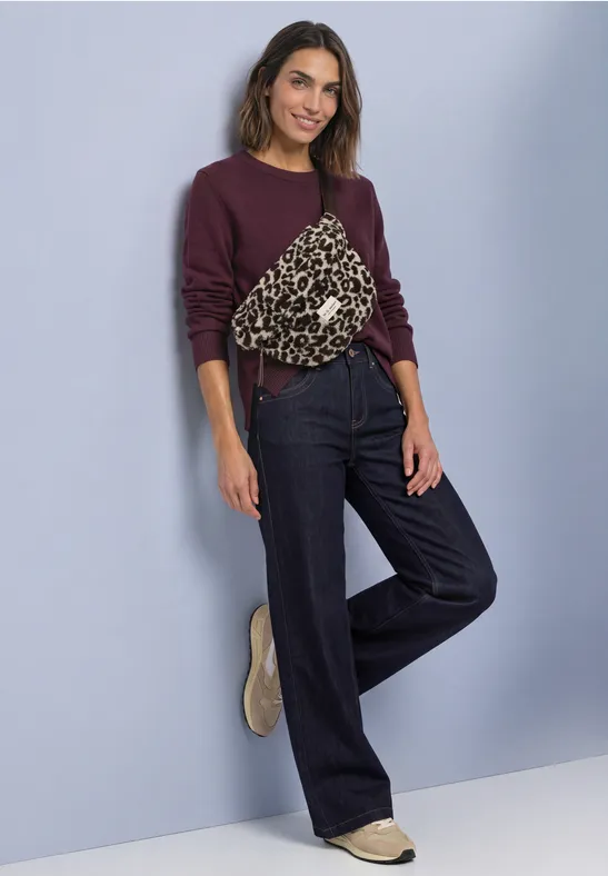 Femme en sweat violet à poche léopard, jean large sombre et chaussures beiges devant un mur bleu clair.