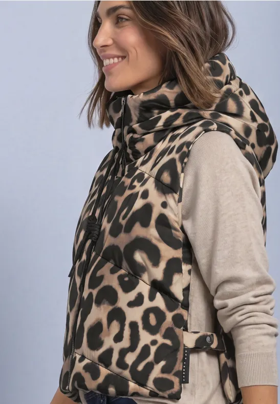 Femme de profil souriante portant une doudoune sans manches à motif léopard à capuche sur un pull beige.