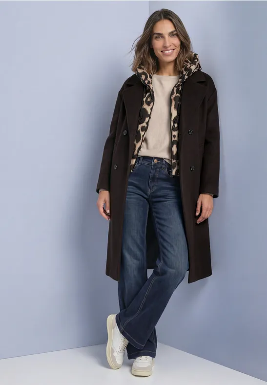 Femme souriante devant un mur clair, manteau brun, pull beige, jean large et baskets blanches.
