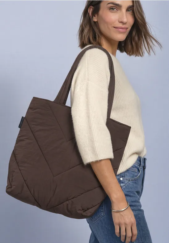 Vrouw in beige trui en blauwe jeans draagt grote, bruine, doorgestikte tote bag over haar schouder.