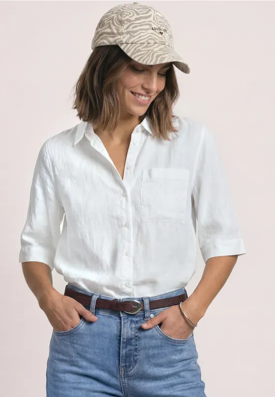 Frau mit weißem Hemd, blauer Jeans und gemusterter Kappe, lächelnd mit Händen in den Taschen.