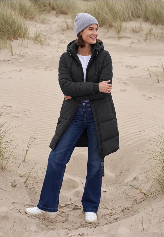 Frau in langer, schwarzer Steppjacke mit Jeans, grauer Mütze und weißen Sneakern steht lächelnd am Dünenstrand.