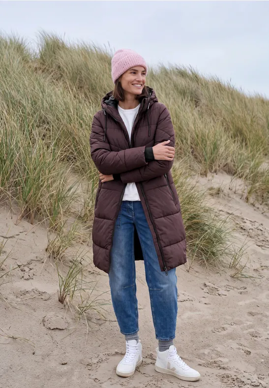 Femme souriante en manteau matelassé, jean et bonnet rose sur les dunes de sable.