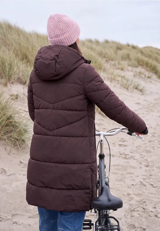 Personne en manteau matelassé marron et bonnet rose pousse un vélo sur des dunes.