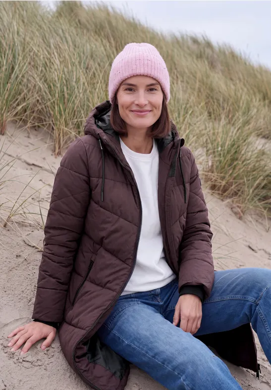 Une femme souriante assise sur la plage près des dunes, vêtue d’une doudoune brune, d’un jean et d’un bonnet rose.