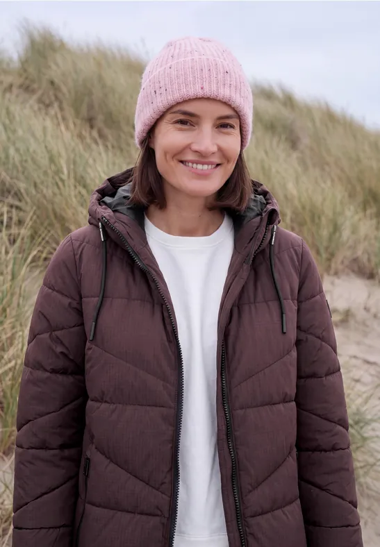 Femme souriante en doudoune marron et bonnet rose devant des dunes herbeuses.